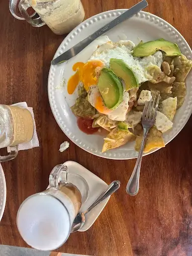 Chilaquiles topped with a runny fried egg, fresh avocado slices, beans, and tangy salsa sit beside coffee drinks for a hearty breakfast at Alquimista