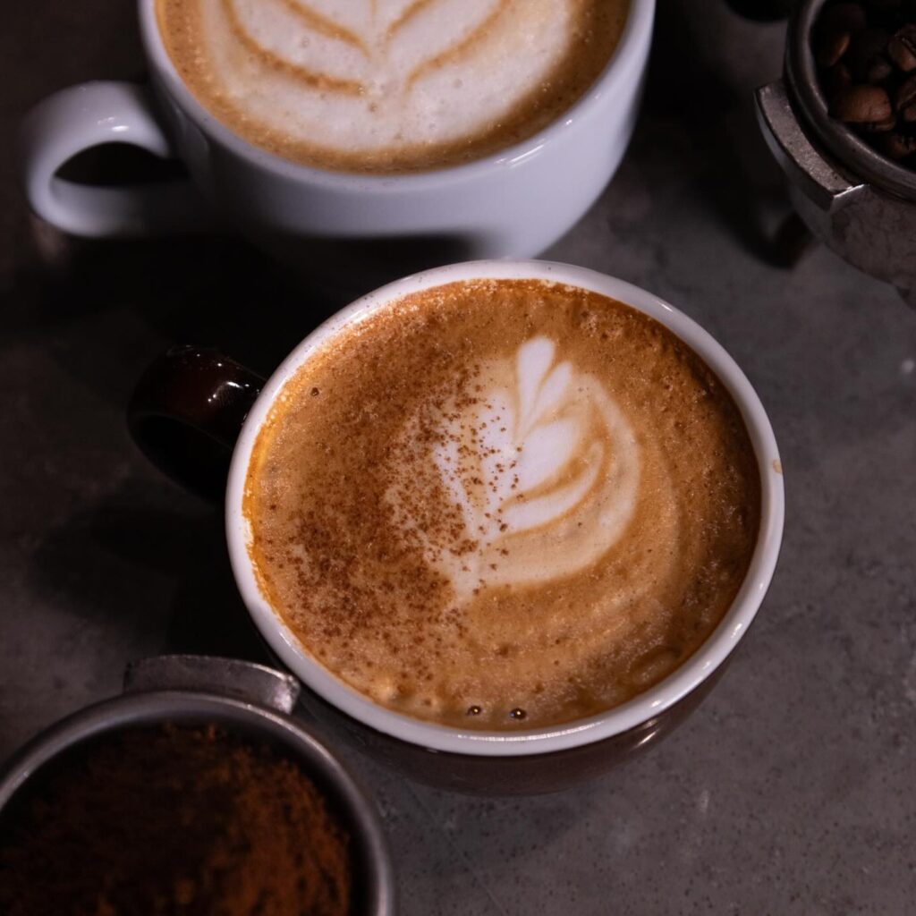 Un cortado caliente se coloca junto a otra taza con café intenso, arte en espuma y un toque de canela que resalta esta preparación de estilo español.