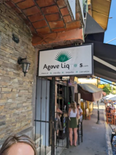 A liquor shop with a white sign hangs over a shaded sidewalk as people stand near the entrance, showing a casual street view in a busy part of town.