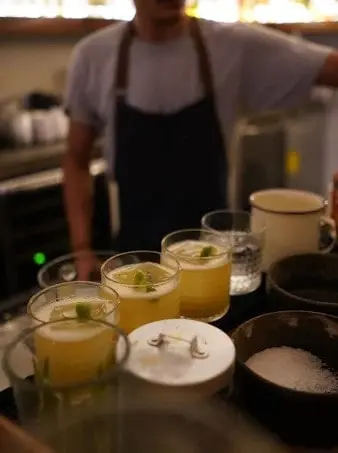Bartender serving several margaritas with lime garnish at a dimly lit Sayulita bar, with cocktail salt, bowls, and bar tools on the counter.