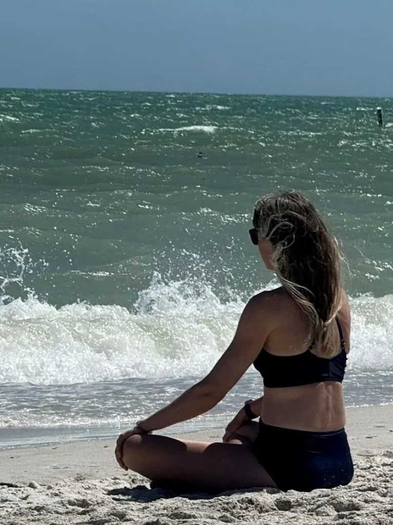 A woman sits in quiet meditation on a Pacific beach at sunset, with rolling waves and open horizon creating a peaceful, restorative shoreline moment.