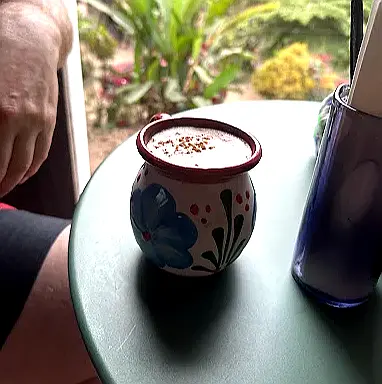 Una taza de cerámica pintada a mano llena de café suave descansa sobre una mesa con vista al jardín en Café Coyote, capturando el ambiente acogedor de este café de Sayulita.