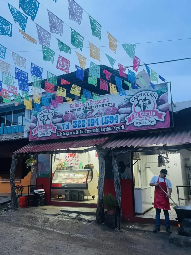 A small butcher shop with colorful papel picado above the entrance is shown at dusk, with a worker preparing food beside the open storefront.