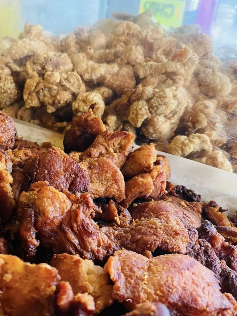 Crispy chicharrón and golden fried pork pieces are piled high behind a display case, showing crunchy texture and rich color in a butcher shop setting.
