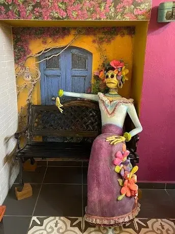 Catrina sculpture seated on a bench in Sayulita with a broken hand, colorful flowers on her dress, and a backdrop reflecting Día de Muertos artistry.