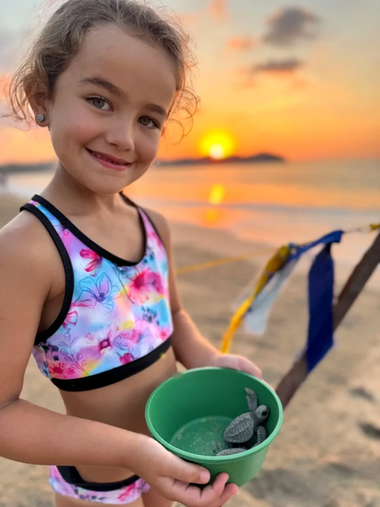 A smiling child stands on the beach at sunset holding a green bucket with two baby sea turtles, with calm ocean water glowing behind her.