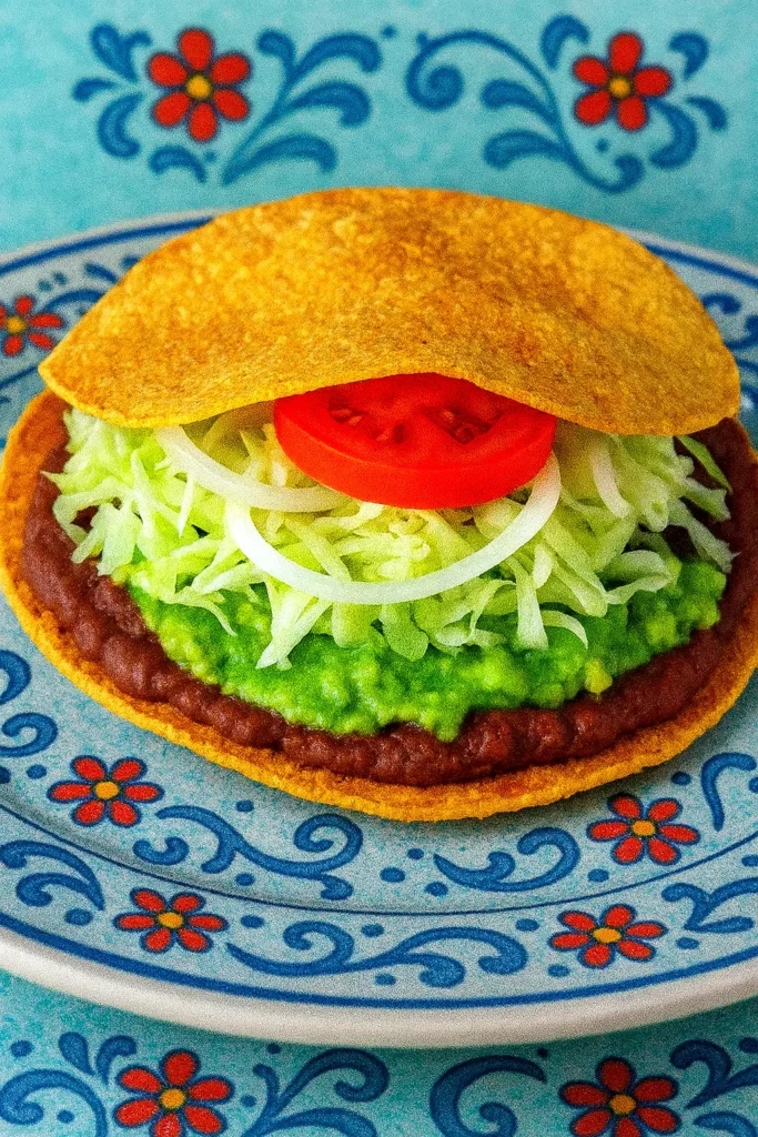 Crisp tostada layered with beans, guacamole, lettuce, onion, and tomato, showcasing fresh textures and bright flavors in a simple Mexican snack.