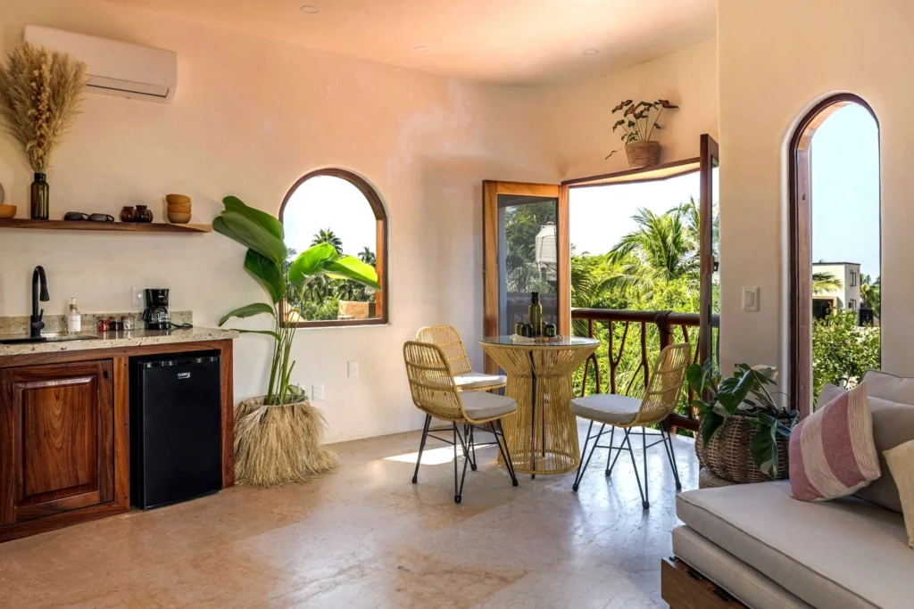 Sunlit open-plan room with kitchenette, café table, lounge seating, and wide terrace doors opening to tropical garden views.