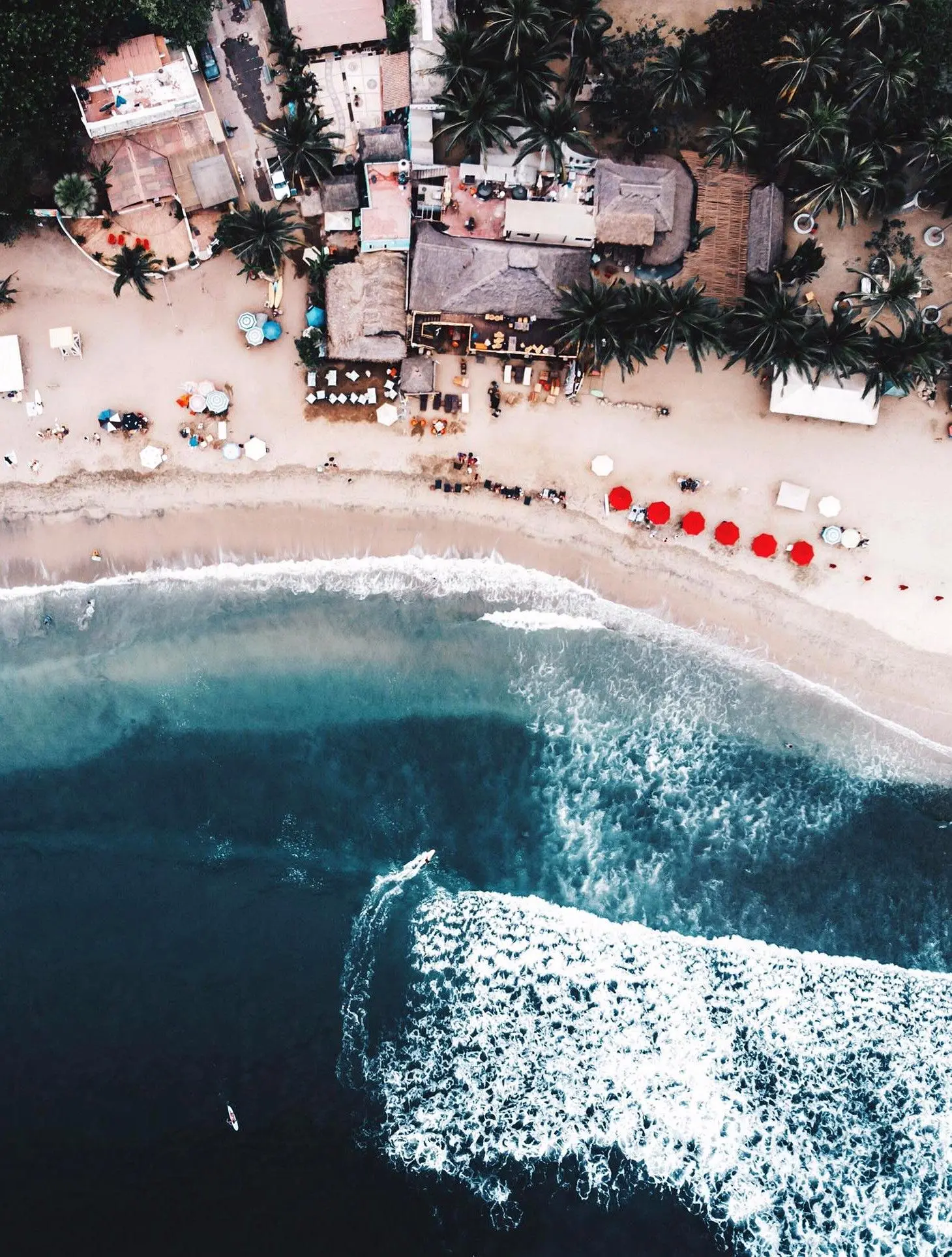 Una vista aérea muestra clubes de playa y mesas mezclándose con la orilla mientras la gente va entre comida y surf.