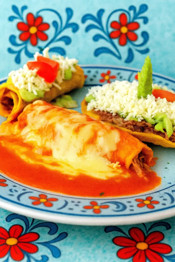 Cheesy red enchilada on a plate beside tacos topped with crema, avocado, and tomato, showing a colorful combination of classic Mexican dishes.