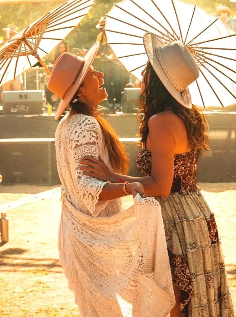Two friends laugh beneath sunlit parasols as music drifts through the festival grounds, capturing a joyful moments at a bright outdoor celebration.