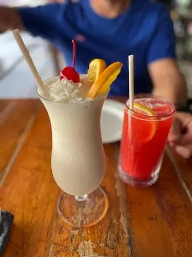 Frozen tropical cocktail with whipped topping, cherry, and citrus garnish beside a bright red drink on a wooden table, served at a bar in Sayulita.