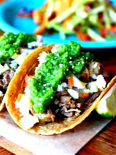 Carnitas tacos topped with onions, cilantro, and salsa verde, served with lime wedges, highlighting tender shredded pork and bright Mexican flavors.