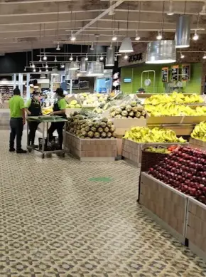 A wide produce section with bananas, apples, pineapples, and other fresh fruit stacked neatly, with staff preparing displays under market lighting.
