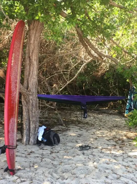 A surfboard rests beside a hammock strung between trees at La Lancha, creating a shaded hideaway where surfers relax between sessions on soft sand.