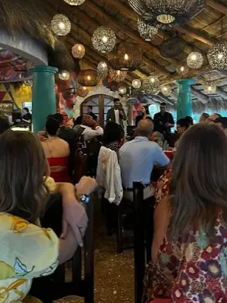 Guests having dinner at a Sayulita restaurant with musicians, colorful décor, woven hanging lights, and a lively setting beneath a thatched roof.