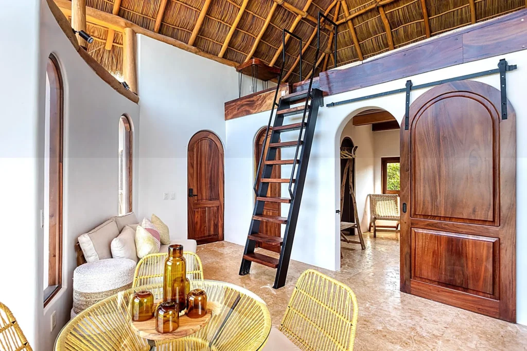 Lofted palapa living space with stairs to the mezzanine, arched wood doors, lounge seating, and dining area inside Amari Boutique Hotel Sayulita.