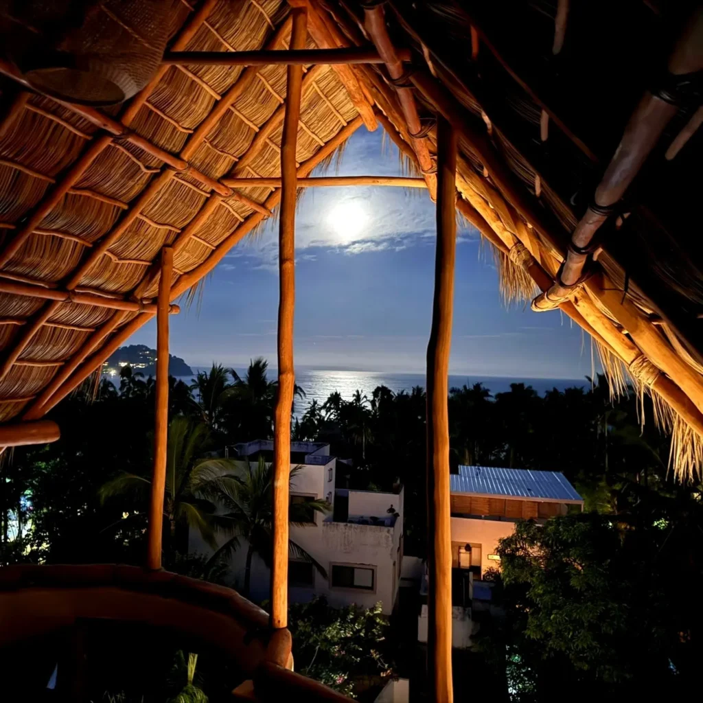 Moonlight reflects over the Pacific from the palapa at the boutique hotel, highlighting handcrafted thatch, warm wood beams tranquil hillside setting.