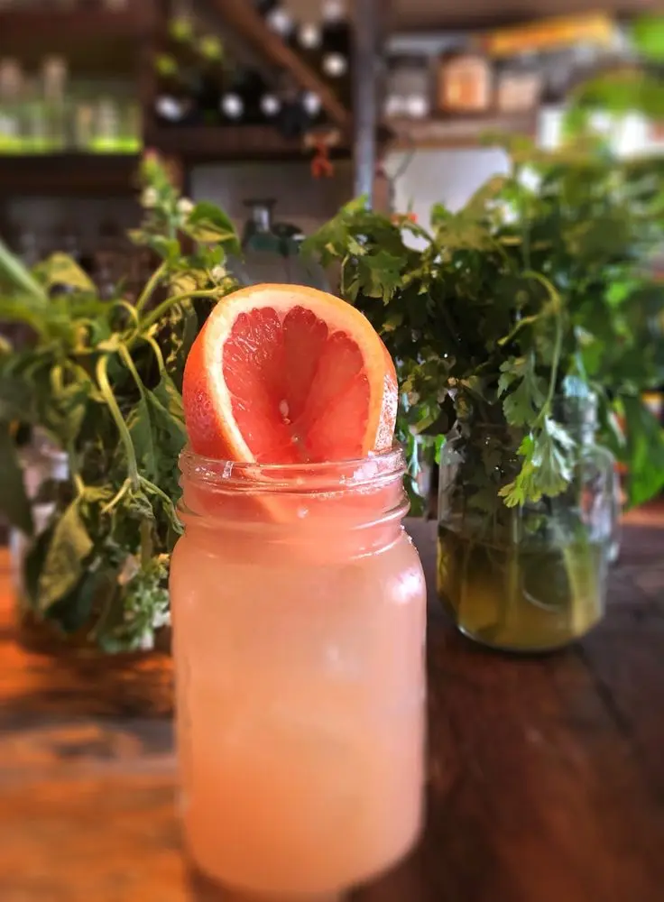 Paloma cocktail in a mason jar topped with a fresh grapefruit slice, with jars of herbs in the background at a rustic bar in Sayulita.