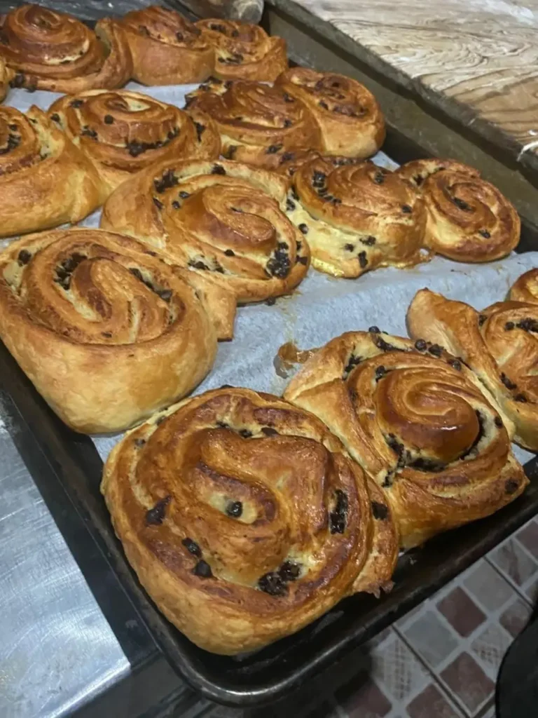 Golden, flaky pastries with swirled layers and chocolate pieces cool on a baking tray, showing crisp texture and fresh-baked detail.
