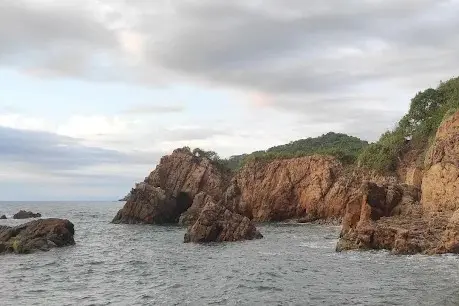 Rust-red cliffs rise above turquoise water where hidden caves open to the sea—a striking vista on the Pacific Riviera Coastal Belt.