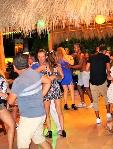 People gather beneath the palapa as lively music and warm lights set a festive mood, creating a joyful night filled with movement and laughter.