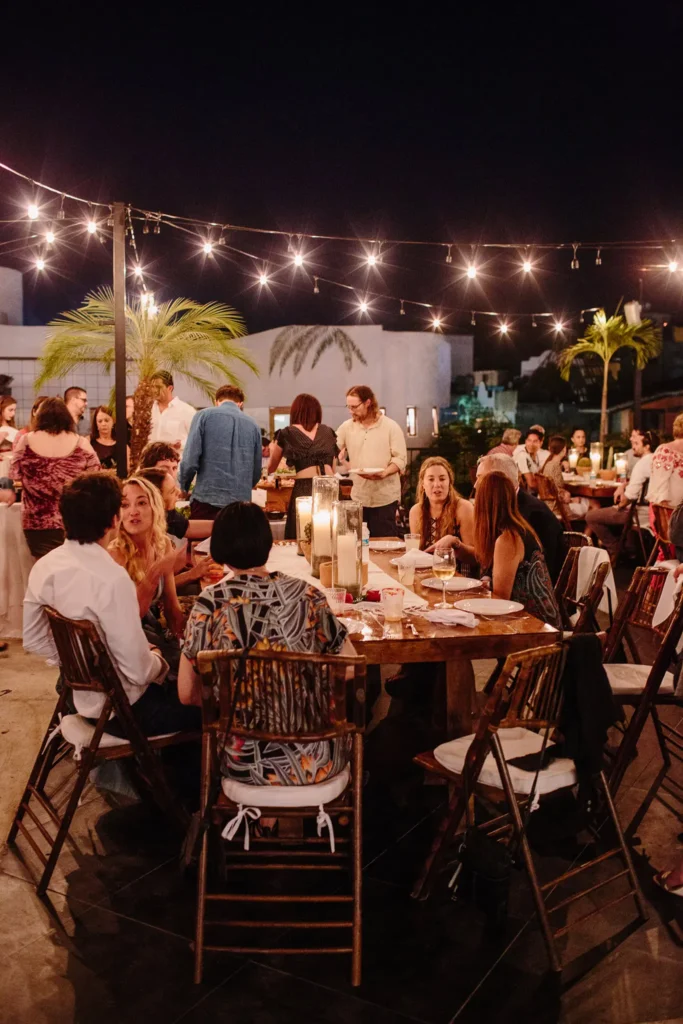 Guests share food and conversation under warm string lights at rustic wooden tables during a relaxed outdoor dinner on a warm evening.