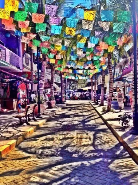 Colorful papel picado flags hang over a lively Sayulita street lined with shops, capturing the vibrant atmosphere of local food and market areas.