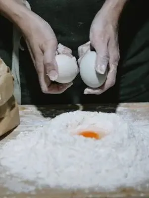 Hands dusted with flour hold two eggs above a mound of flour with an egg yolk in the center, capturing the start of a home-cooked meal in Sayulita.