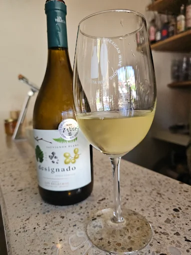 A glass of chilled white wine sits on a countertop beside its bottle, with shelves and barware behind it in a small wine shop.