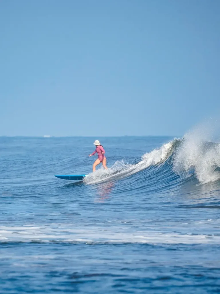 A surfer in a pink rash guard glides gracefully along a mellow Pacific wave beneath a clear blue sky, capturing the calm rhythm of the Riviera Nayarit coast.