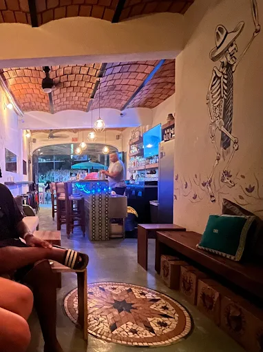 Blue-lit bar in Sayulita with a bartender preparing drinks beside a playful skeleton mural, with streetside seating in a casual setting.