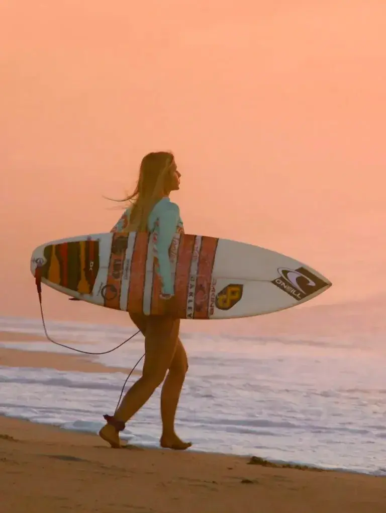 Surfer carries board into sunrise waves near her wellbeing hotel — a passionate hideaway of ocean rhythm and unhurried energy.