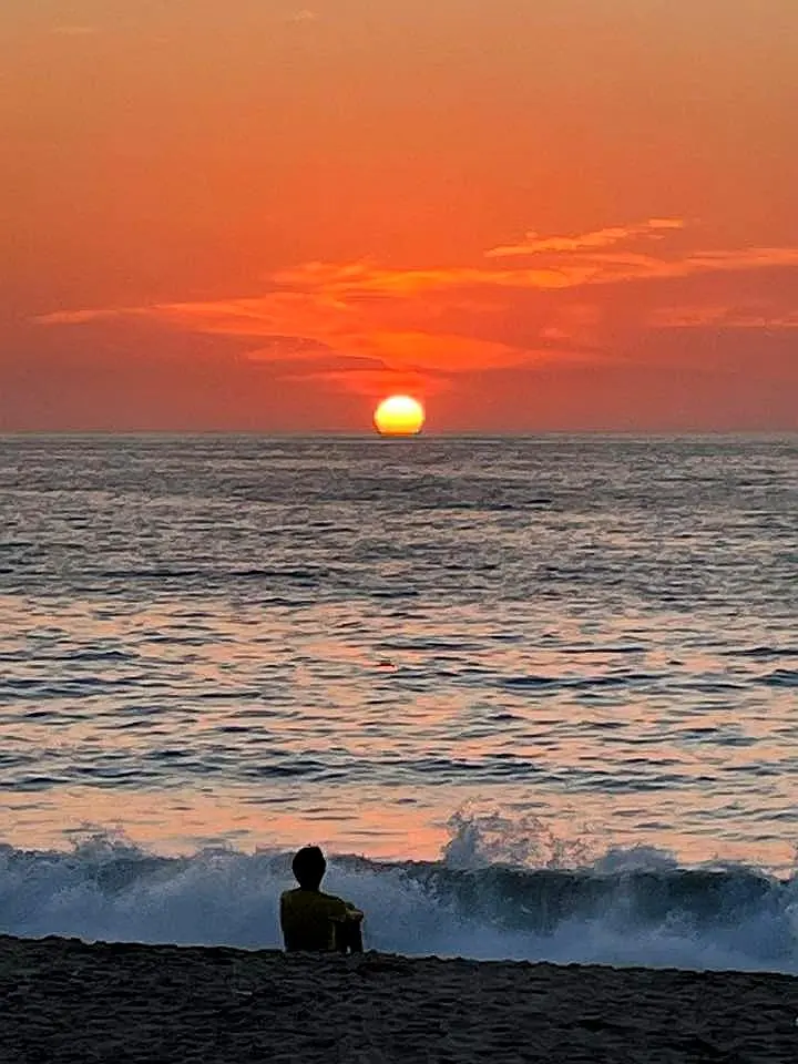 A warm sunset settles over gentle waves as a lone beachgoer watches the horizon, creating a calm shoreline moment with a soft coastal retreat feel.