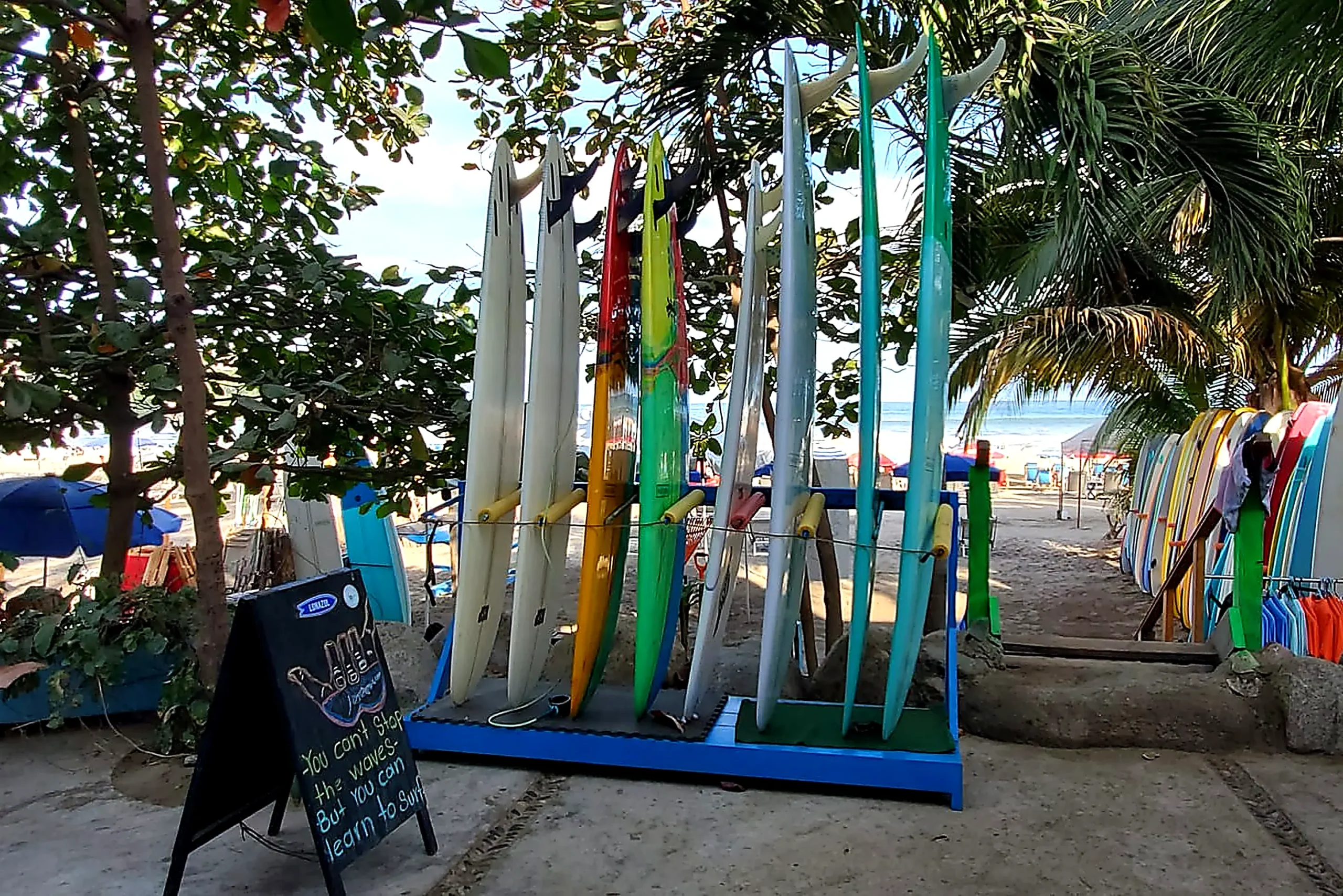 Colorful surfboards lined up near the sand in Bahía de Banderas—palms overhead, ocean views ahead, classic surf vibe.
