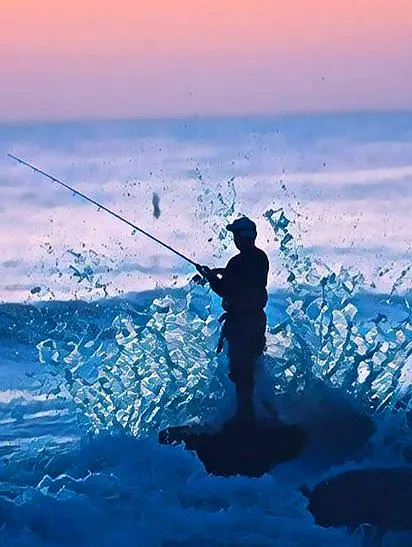 A silhouetted angler casts into glowing waves at dusk, adding a peaceful fishing moment to a relaxed oceanfront retreat.