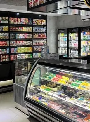 A refrigerated deli case filled with cheeses and packaged foods stands beside shelves stocked with canned and bottled items in a small grocery store.