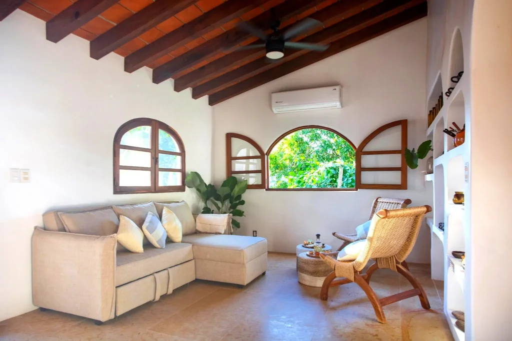 Bright Villa Ama living room with arched windows, plush sectional, rattan chairs, terracotta beams, and garden views in Sayulita, Nayarit.
