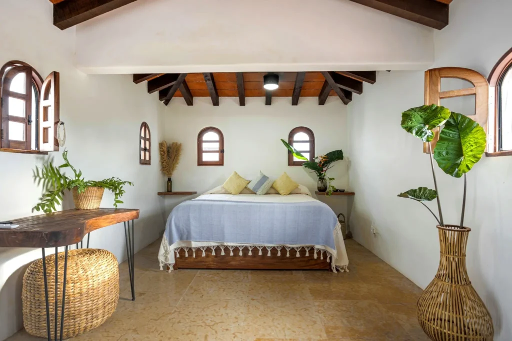 Studio bedroom with arched windows, terracotta beams, queen bed and blue throw, styled with plants and warm light inside Amari in Bahía de Banderas.