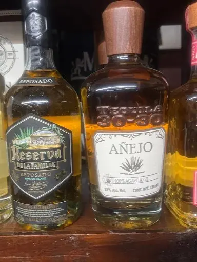 A row of tequila bottles, including añejo and reposado varieties, displayed on a wooden shelf with distinct labels, glass shapes, and amber tones.