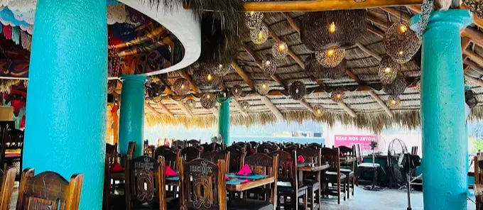 Open-air dining at Xochi with artisan wooden chairs, woven lantern lighting, turquoise columns, and a palapa roof, creating an elevated coastal seafood atmosphere.