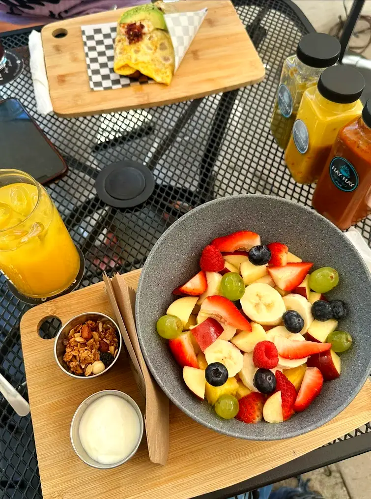 Colorful fruit salad with bananas, strawberries, grapes, granola, and yogurt on a wooden board beside orange juice and a breakfast wrap at YAH-YAH