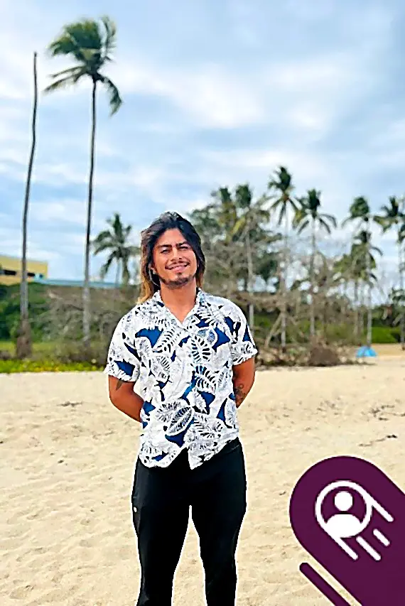 A man smiles on a sandy beach with palm trees behind him, wearing a patterned short-sleeve shirt and black pants under a cloudy sky.