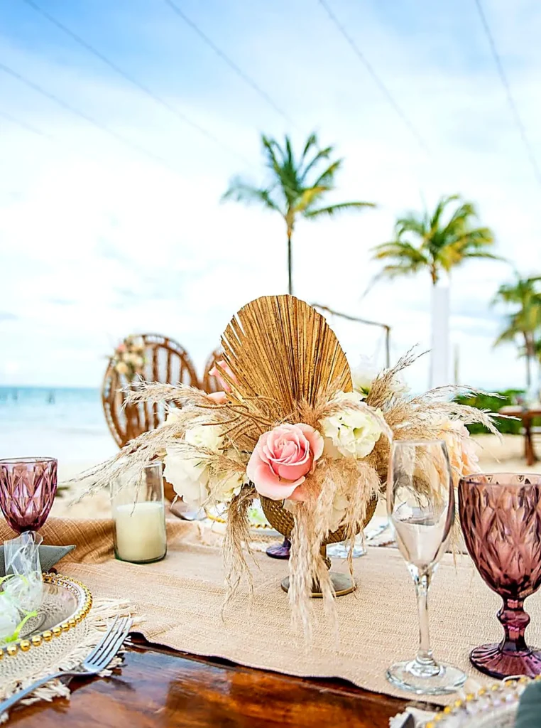 A styled wedding table with floral centerpiece and glassware sits on the sand with palms and ocean, set for an intimate Riviera Nayarit gathering.