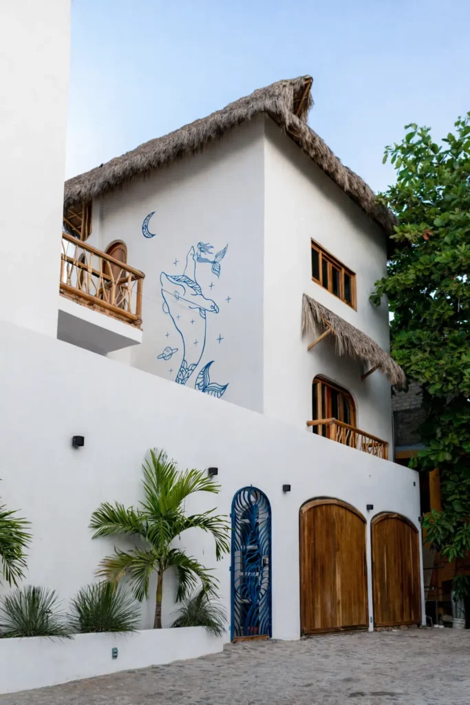Exterior blanco con techo de palapa de palma, puertas de madera tallada, plantas tropicales y un mural inspirado en el océano en Amari Boutique Hotel en Sayulita.