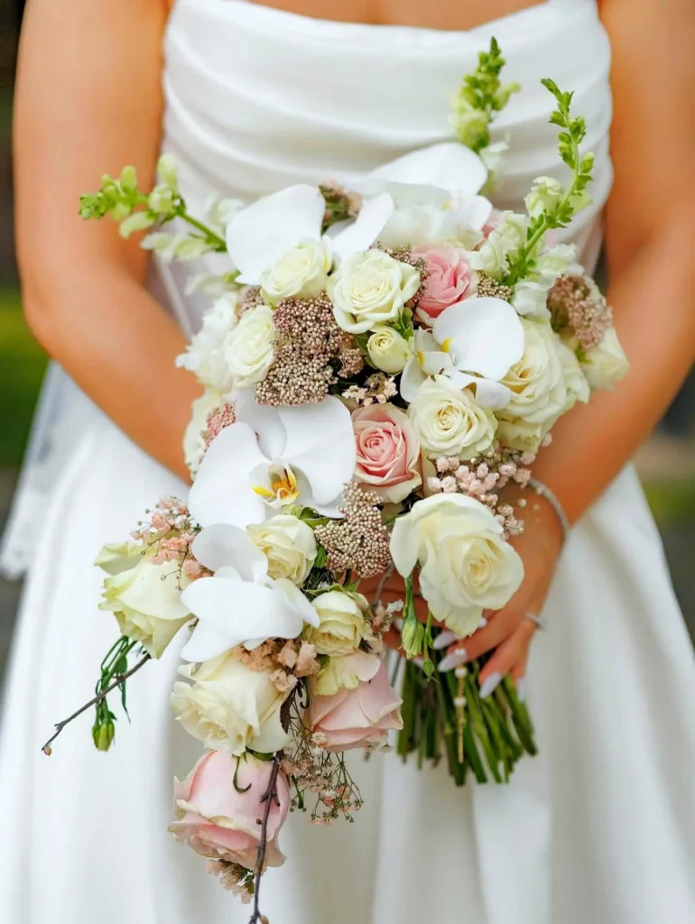 A bride holds a cascading bouquet of white orchids, blush roses, and delicate florals against a simple gown, highlighting softness and calm elegance.