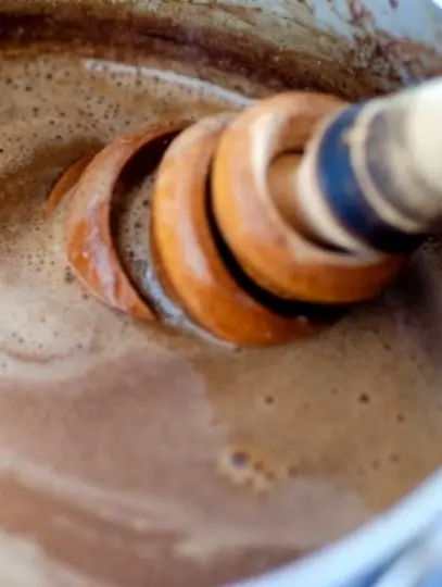 A wooden whisk stirs thick champurrado in a pot, showing rich texture and slow blending that creates deep cocoa richness.
