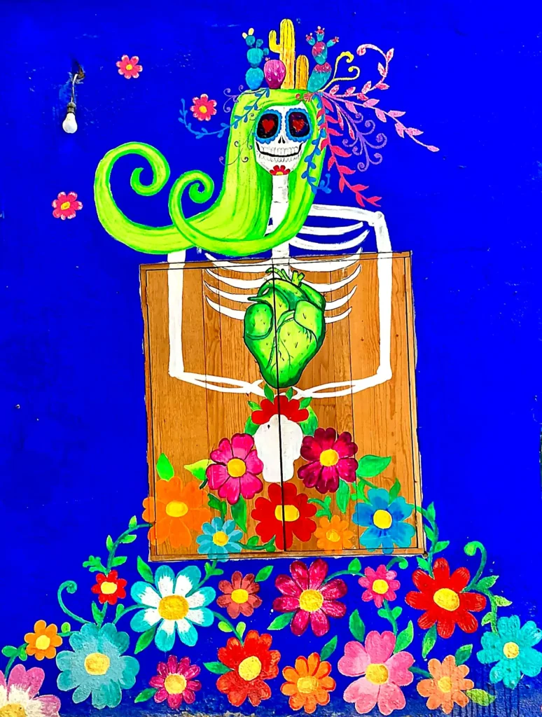 Vibrant Día de los Muertos mural shows a smiling skeleton with green hair and blooming flowers on a bright blue wall along Bahía de Banderas.