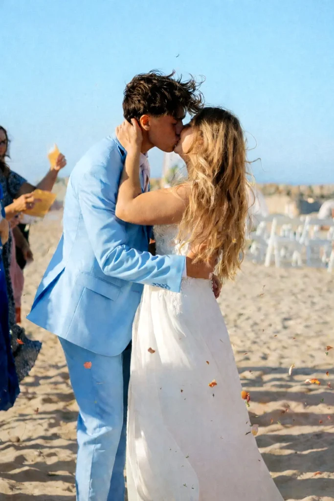 A newly married couple shares their first kiss on the sand as petals fall around them, capturing joy and movement during an outdoor wedding ceremony.