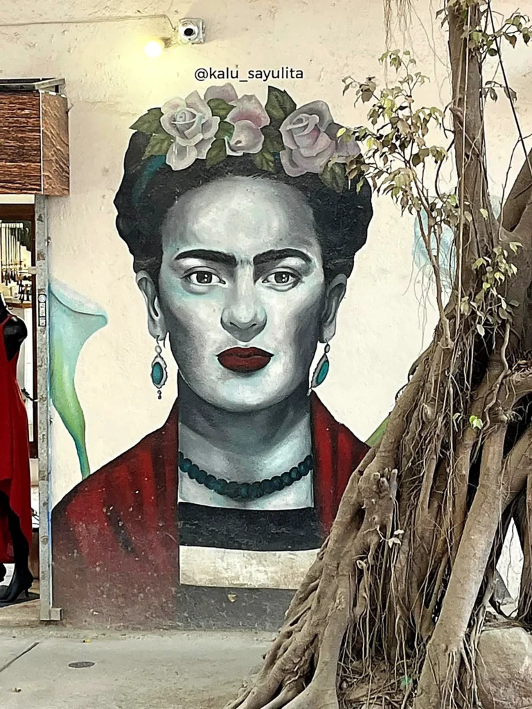 Frida Kahlo mural with floral crown and red shawl painted beside sprawling banyan roots, capturing a quiet, iconic scene.
